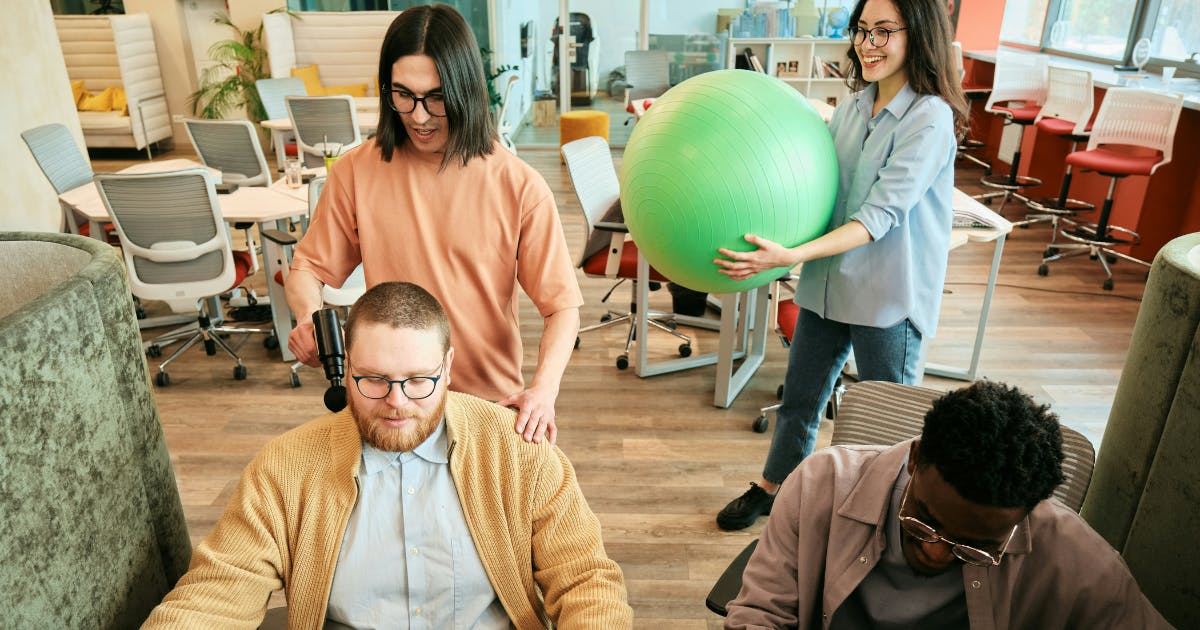 Employees enjoying a break and participating in wellbeing activities, using a massage gun and yoga ball to promote physical and mental health in the workplace.